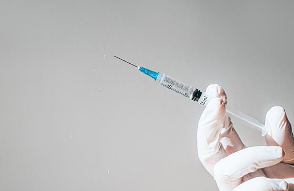 Imagem colorida de uma mão usando luvas segurando uma seringa ára aplicação de vacina