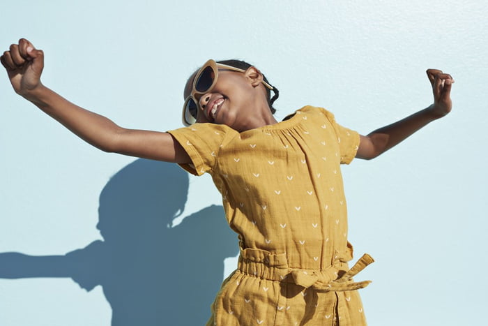 Criança pulando em frente a parede azul. Ela está usando óculos de sol e com os braços abertos