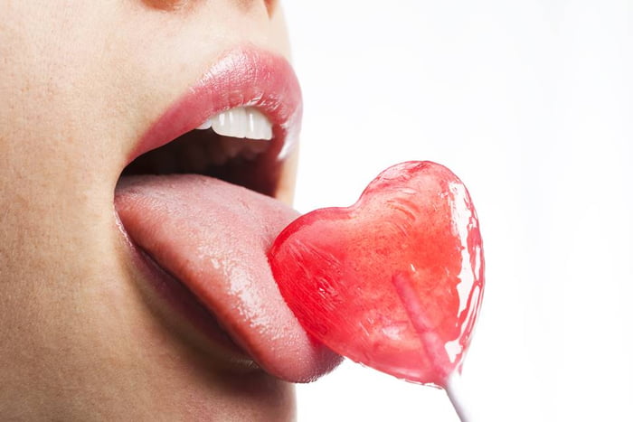 Foto colorida em close da boca de uma mulher lambendo um pirulito de coração - Metrópoles