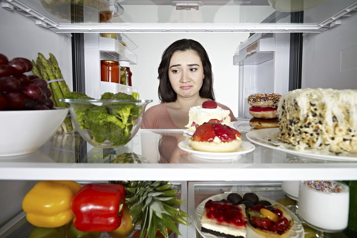 Foto colorida de mulher branca com a cabeça dentro da geladeira, cheia de bolos, doces, legumes e frutas - Metrópoles