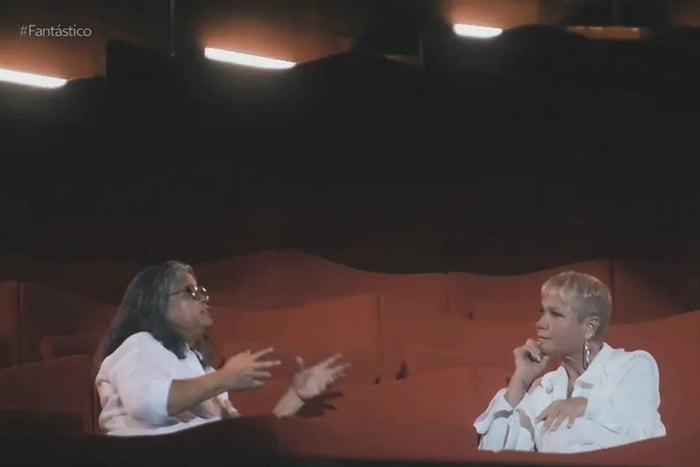 xuxa sentada em poltrona de sala de teatro conversando com Marlene Mattos, uma mulher de cabelos cinzas e óculos. As duas vestem branco - metrópoles