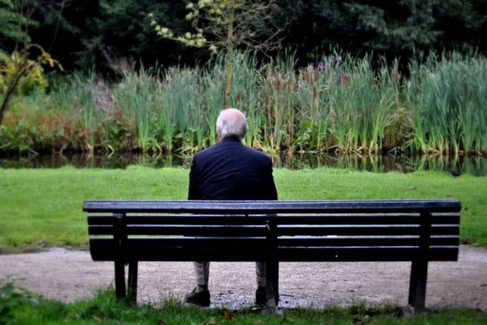 Imagem colorida de idoso sentado sozinho em um banco, em um parque - Metrópoles
