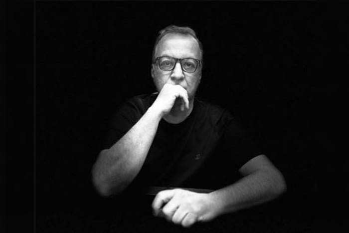 Imagem em preto e branco de Carlinhos Cachoeira, de óculos, sentado, com uma mão sobre a mesa e outra embaixo do queixo