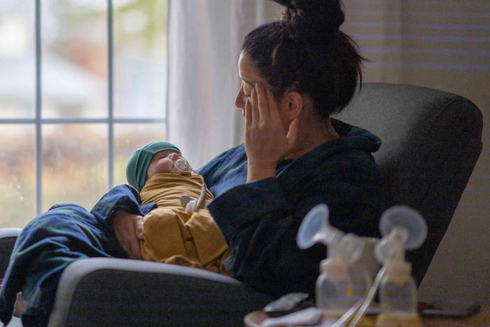 Foto mostra mãe com bebê em seu colo. Ela tampa os olhos com a mão - Metrópoles - depressão pós-parto