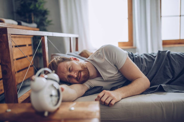 Foto mostra homem deitado em cama e aproximando o braço de um despertador - Dormir demais - Metrópoles