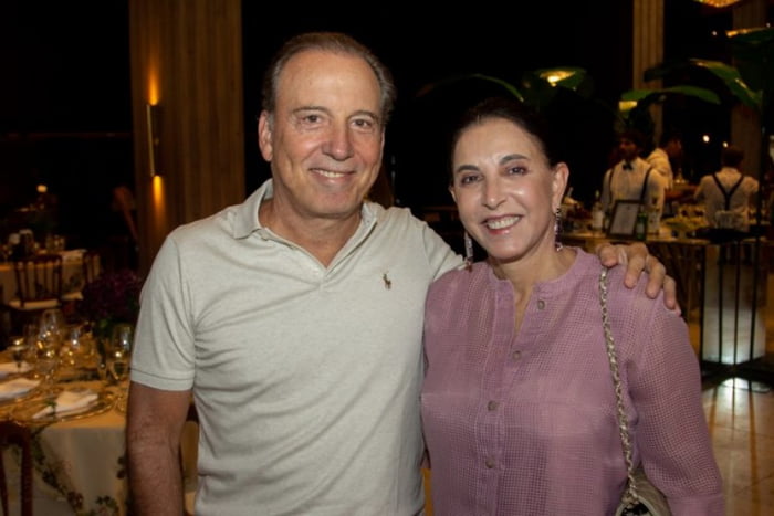 Imagem colorida mostra casal sorrindo para foto; o homem, o banqueiro Binho Bezerra, idoso, veste camiseta branca, a mulher veste roupa rosa. Ambos foram encontrados mortos no Guarujá - Metrópoles