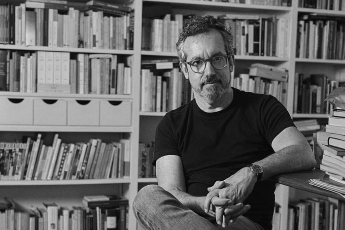 Imagem em preto e branco de homem com camisa preta de óculos sentado em frente a livros - Metrópoles
