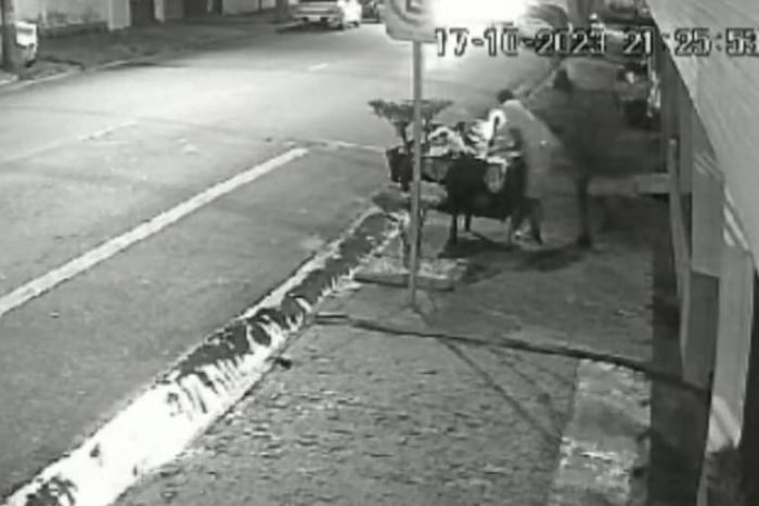 Imagem em preto e branco mostra frame de vídeo de câmera de segurança com um homem atacando outro com facadas enquanto ele mexe em lixeira de rua - metrópoles