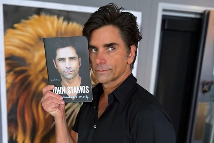 Foto colorida de um homem branco, com cabelo preto e blusa preta segurando um livro - Metrópoles