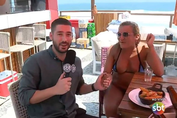 foto colorida de homem com microfone conversando com mulher loira que usa biquini. Os dois estão sentados em cadeiras na orla da praia - metrópoles