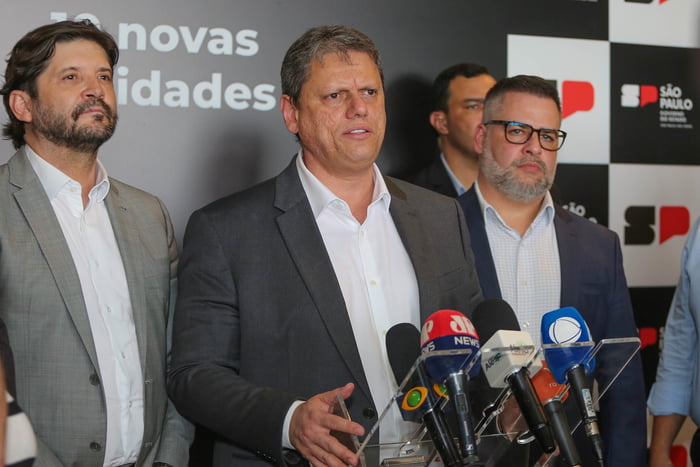 Imagem colorida mostra o governador Tarcísio de Freitas, homem branco, grisalho, de terno cinza e camisa branca, falando em um púlpito repleto de microfones ao lado de aliados políticos