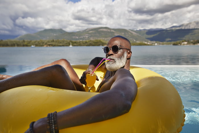 Homem careca idoso negro usando óculos escuros enquanto bebe suco em anel inflável durante férias - Metrópoles