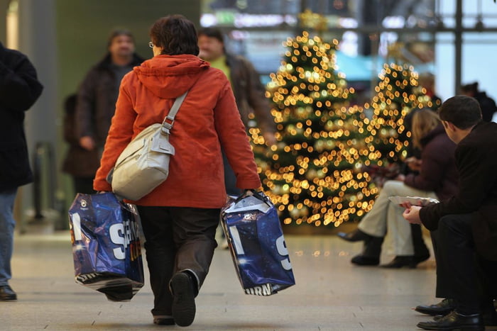 Mulher faz compras em shopping na época do Natal. Ela carrega sacolas e veste um casaco vermelho, aparecendo de costas na imagem. Ao fundo, uma árvore de Natal - Metrópoles