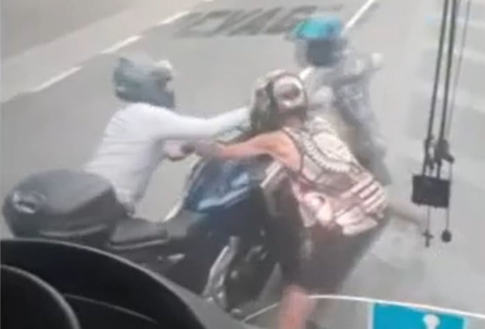 Imagem de dois assaltantes de capacete tentando pegar moto de motociclista em avenida - Metrópoles