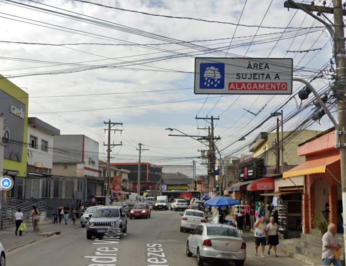 Imagem mostra rua, com carros e placa de sinalização de área sujeita a alagamentos - Metrópoles