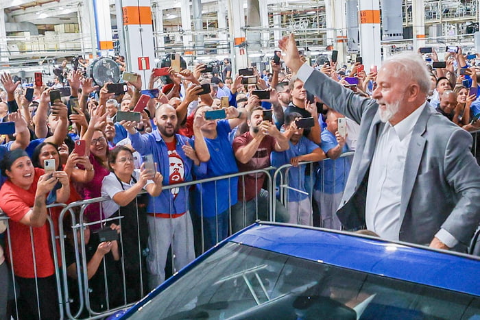 Imagem colorida mostra Lula em pé em um carro conversível, acenando para um grupo de operários que sorriem ao vê-lo
