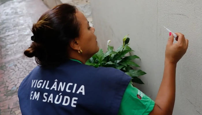 Imagem de mulher com colete da Vigilância Sanitária olhando para frasco de coleta - Metrópoles
