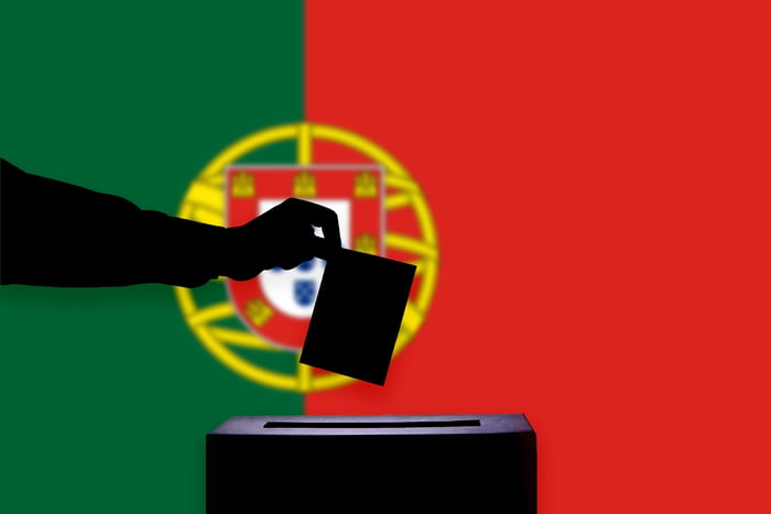 Imagem colorida mostra bandeira de Portugal ao fundo, com uma mão, contra a luz, depositando um voto em uma urna - Metrópoles