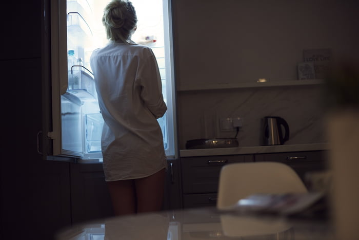 Foto colorida de uma mulher de camisa branca de costas olhando a geladeira na cozinha escura - Metrópoles