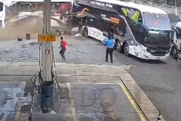 Imagem congelada de vídeo de acidente em posto de combustível. Um ônibus atingido na traseira, uma pessoa de camisa azul observa e outra pessoa de camiseta vermelha, corre.